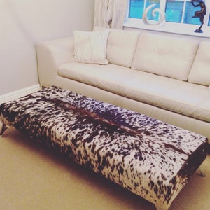 May include: A black and white cowhide ottoman with a white leather sofa in the background.