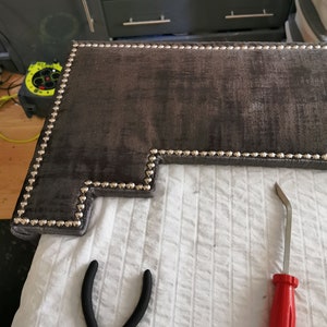 May include: A close-up of a dark gray headboard with silver upholstery tacks. A red tool and black pliers are on a white surface.
