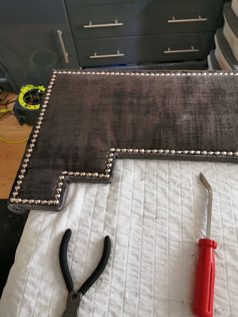 May include: A close-up of a dark gray headboard with silver upholstery tacks. A red tool and black pliers are on a white surface.