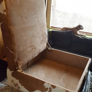 May include: A brown and white cowhide storage ottoman with a hinged lid. The lid is open and the inside is lined with a light brown fabric.