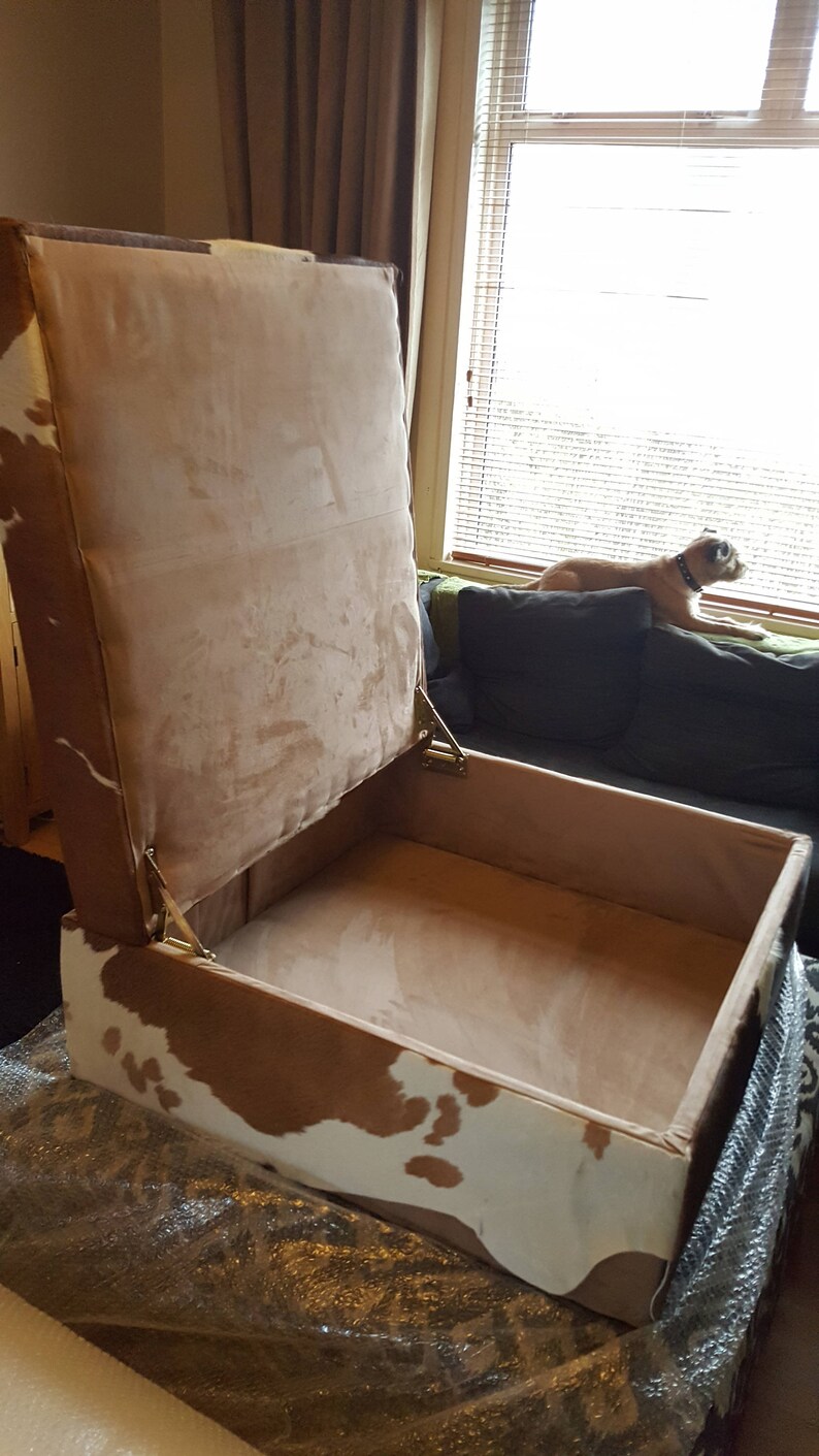May include: A brown and white cowhide storage ottoman with a hinged lid. The lid is open and the inside is lined with a light brown fabric.