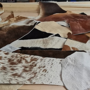 May include: A collection of assorted cowhide leather scraps in various shapes and sizes. The pieces feature natural patterns and textures in shades of brown, white, black, and tan, with some sections displaying spotted or solid color variations.