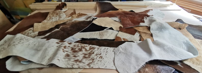 May include: A collection of assorted cowhide leather scraps in various shapes and sizes. The pieces feature natural patterns and textures in shades of brown, white, black, and tan, with some sections displaying spotted or solid color variations.
