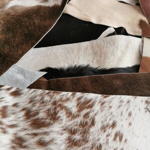 May include: A collection of overlapping cowhide leather scraps in various patterns and colors, including solid black, solid brown, white, and brown-and-white speckled hides. The pieces show natural hair texture and irregular, raw-cut edges.