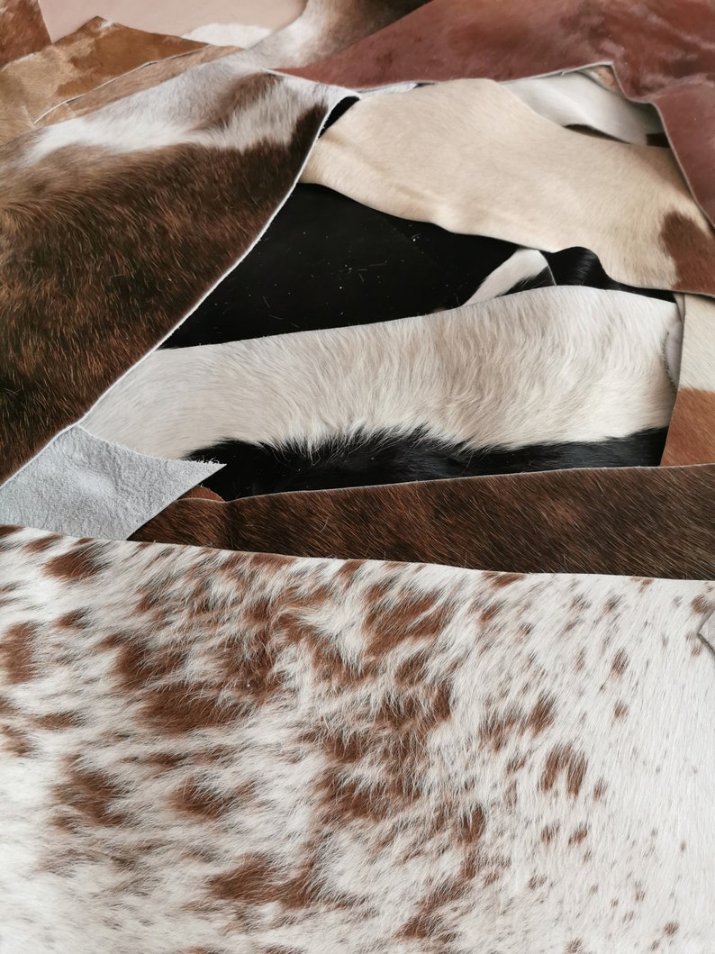 May include: A collection of overlapping cowhide leather scraps in various patterns and colors, including solid black, solid brown, white, and brown-and-white speckled hides. The pieces show natural hair texture and irregular, raw-cut edges.