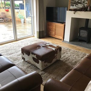 May include: A living room with a brown leather sofa, a brown leather armchair, and a cowhide ottoman. The room has a large window with a view of a patio and a swimming pool. There is a fireplace with a wood-burning stove and a television on the wall.