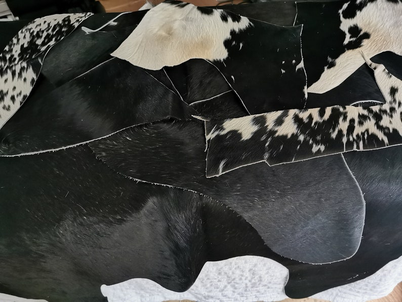May include: A collection of irregular cowhide leather scraps featuring a mix of solid black pieces and black and white spotted patterns. The pieces have a natural hair-on texture with varying shapes and sizes, showcasing the organic hide edges.