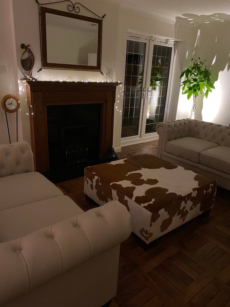 May include: A white couch with a brown and white cowhide ottoman in front of a fireplace with a string of lights. The room has a wooden floor and a large window with a view of a garden.