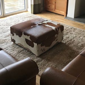 May include: A brown and white cowhide ottoman with a storage compartment sits on a beige shag rug in a living room. Two brown leather sofas are in the background.