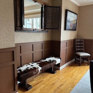 May include: A brown leather bench with a white and brown cowhide seat cushion. The bench is in a dining room with wood floors and brown wainscoting.