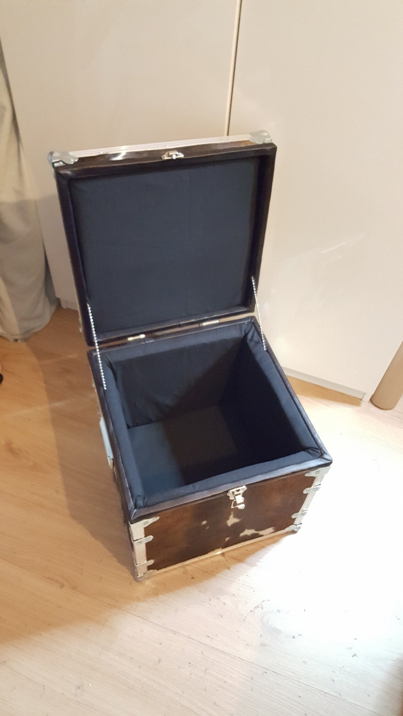 May include: A brown and white cowhide storage trunk with silver metal accents and a black fabric lining. The trunk is open and the lid is propped up.