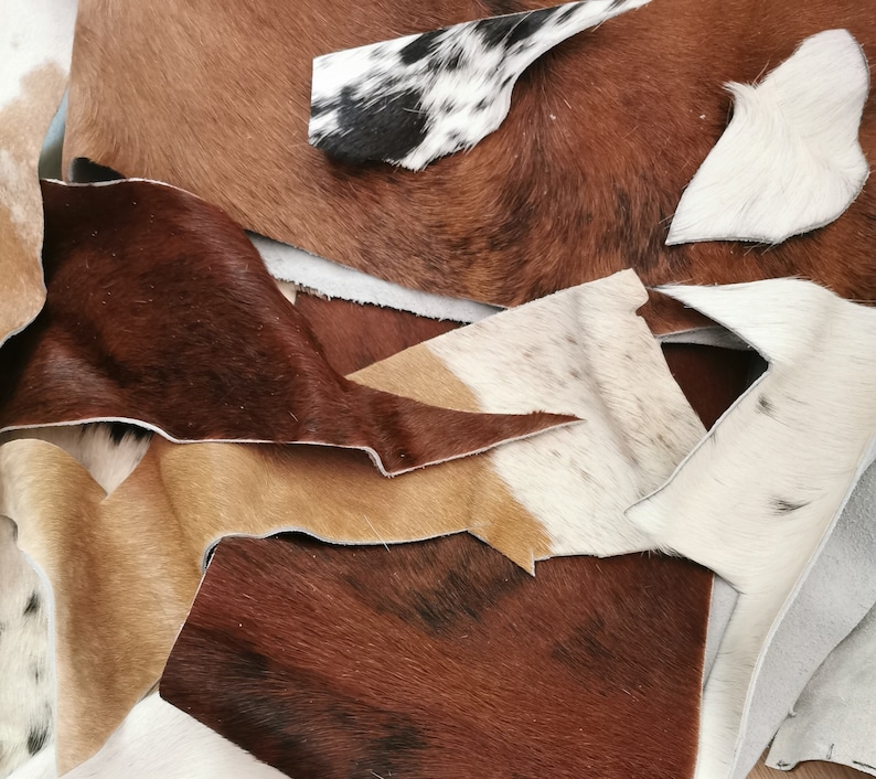 May include: A collection of assorted cowhide leather scraps featuring various natural patterns and textures. The pieces display a mix of solid brown, tan, white, and black-and-white spotted fur, with visible raw edges and irregular shapes.