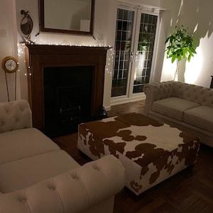 May include: A living room with a white couch, a white loveseat, a brown and white cowhide ottoman, a fireplace with a black firebox, and a potted plant in the corner.
