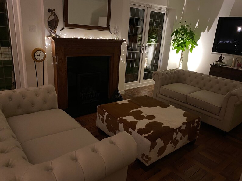 May include: A living room with a white couch, a white loveseat, a brown and white cowhide ottoman, a fireplace with a black firebox, and a potted plant in the corner.