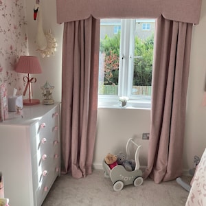 This picture shows a little girls bedroom with a pink and white coloured scheme.  

There is a large window which has a pink pelmet with a shaped bottom and matching floor length lined curtains.