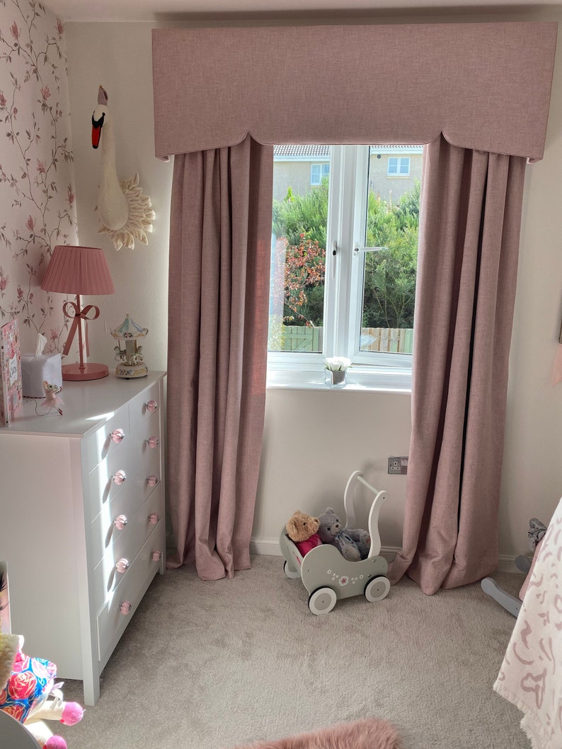 This picture shows a little girls bedroom with a pink and white coloured scheme.  

There is a large window which has a pink pelmet with a shaped bottom and matching floor length lined curtains.