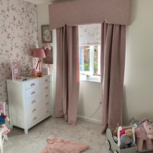May include: A pink and white floral wallpapered room with a white dresser, a pink rug, and pink curtains with a valance. A window with a white roller blind with a floral pattern is visible. A toy stroller and a rocking horse are on the floor.