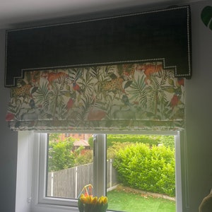 May include: A black and white window blind with a tropical print featuring tigers, leopards, birds, and palm leaves. The blind is raised to reveal a view of a backyard with green grass and a fence.