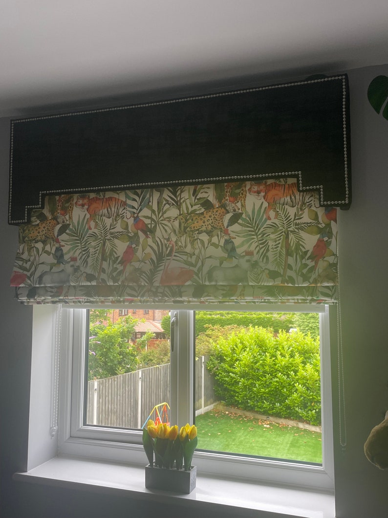 May include: A black and white window blind with a tropical print featuring tigers, leopards, birds, and palm leaves. The blind is raised to reveal a view of a backyard with green grass and a fence.