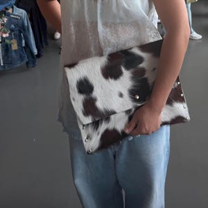 May include: A rectangular clutch bag featuring a cowhide pattern with dark brown and white fur texture. The bag has a fold-over flap design and is accented with small, round silver-toned metal studs along the bottom and side edges of the exterior.