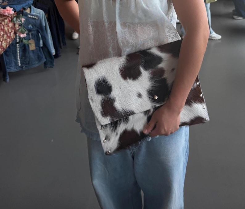 May include: A rectangular clutch bag featuring a cowhide pattern with dark brown and white fur texture. The bag has a fold-over flap design and is accented with small, round silver-toned metal studs along the bottom and side edges of the exterior.