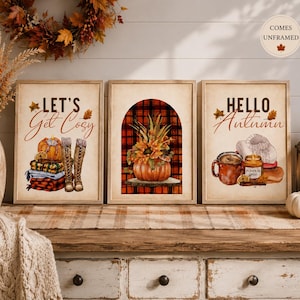 May include: Three rectangular wall art prints featuring autumn themes. The left print shows a pumpkin, folded blankets, and boots with the text "LET'S Get Cosy". The centre print displays a pumpkin with flowers inside an arch over a red plaid background. The right print shows a mug, a candle labelled "PUMPKIN spice", and a sweater with the text "HELLO Autumn".