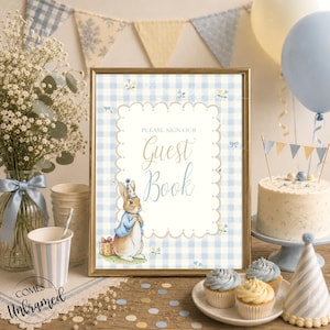 May include: A rectangular sign in a gold frame featuring a light blue and white gingham background with a scalloped white centre panel. The text reads Please Sign Our Guest Book above an illustration of a rabbit wearing a blue jacket and party hat.