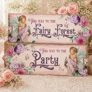 May include: Two rectangular wooden arrow signs with a light wood grain finish. The top sign reads "This way to the Fairy Forest" and the bottom sign reads "This way to the Party". Both feature illustrations of a fairy and purple and pink flowers.