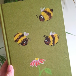 Op de afbeelding: Een groen notitieboek met handgeschilderde illustraties van bijen en een bloem. De bijen zijn geel en bruin met kleine roze vleugels. De bloem is roze en rood met een gele kern en groene stengel.