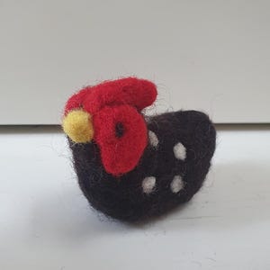Può includere: Figurina di gallina in feltro fatta a mano. La gallina è nera con pois bianchi e una cresta e bargigli rossi. Una piccola palla gialla è sulla testa della gallina. La figurina è una decorazione stravagante.
