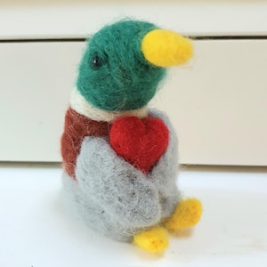 Op de afbeelding: Een naaldgevilte wilde eend-figuur met een groene kop, gele snavel, witte nekring, bruine borst en grijze vleugels. De eend houdt een klein, felrood hartje tegen de borst met de vleugels en heeft gele voeten.