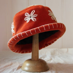 May include: A bright orange felt hat with white embroidered flowers. The hat has a white stitched brim and is displayed on a wooden stand.