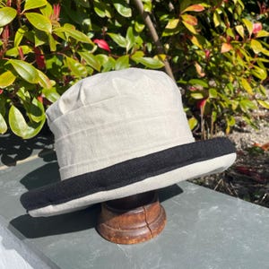 Peut inclure: Un chapeau en lin blanc avec une bande noire autour du bord. Le chapeau est posé sur un support en bois.