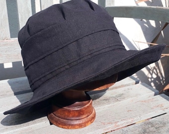 May include: A black linen sun hat with a wide brim and a folded crown. The hat is sitting on a wooden display stand.
