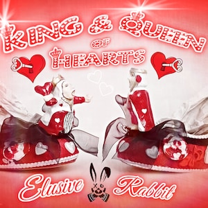 May include: A pair of red high-heeled shoes with a white and black design. The shoes are decorated with hearts, keys, and a queen and king of hearts. The text "King & Queen of Hearts" is written above the shoes. The text "Elusive Rabbit" is written below the shoes.