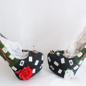 May include: A pair of black high-heeled shoes with a green and red playing card design. The shoes have a white lace bow on one shoe and a red ribbon bow on the other.