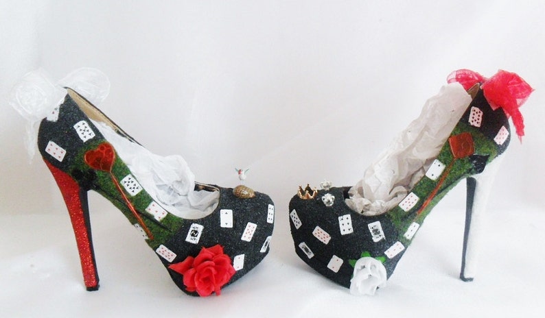 May include: A pair of black high-heeled shoes with a green and red playing card design. The shoes have a white lace bow on one shoe and a red ribbon bow on the other.