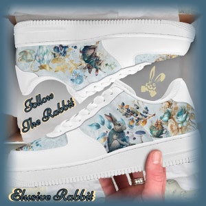 May include: White low-top sneakers with a blue and white floral print featuring a rabbit and a teacup. The text "Follow The Rabbit" is printed on the side of the shoe.