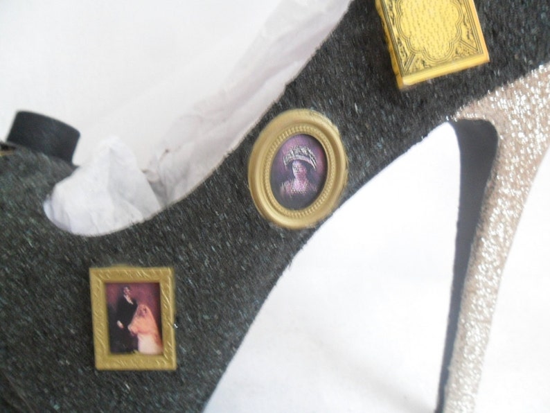May include: A black high-heeled shoe with gold accents. The shoe is decorated with three small framed pictures. One picture features a woman in a crown, another picture features a couple, and the third picture features a decorative design.