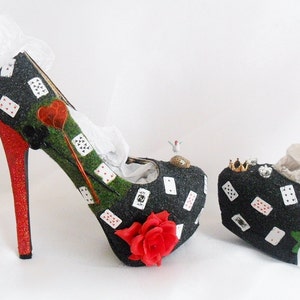 May include: A pair of black high-heeled shoes decorated with playing cards, green grass, and a red rose. The shoes have a red glitter heel and a white lace trim.