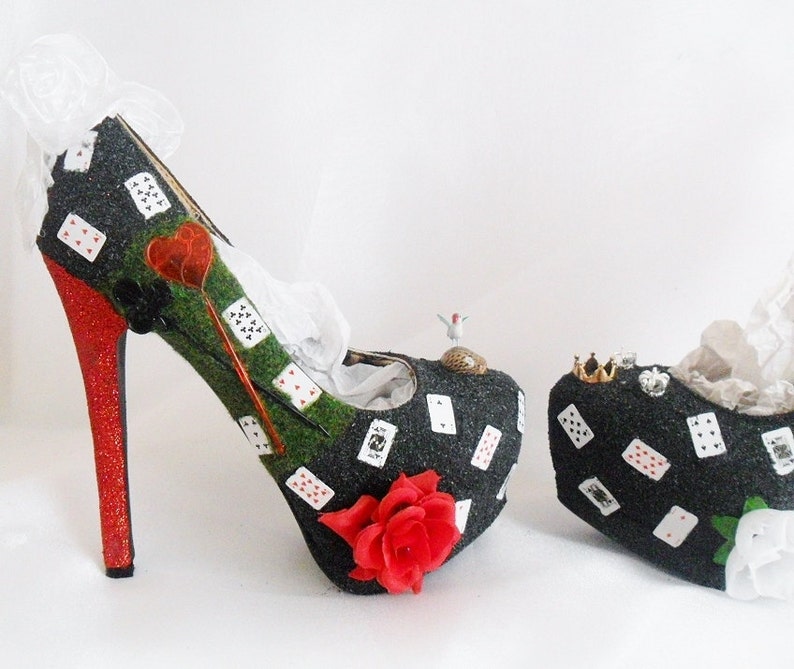 May include: A pair of black high-heeled shoes decorated with playing cards, green grass, and a red rose. The shoes have a red glitter heel and a white lace trim.