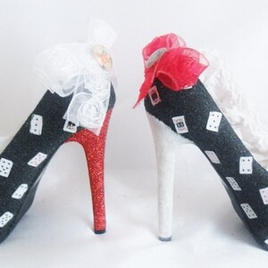 May include: A pair of black high-heeled shoes with white playing card designs. One shoe has a red glitter heel and a white bow, while the other has a white heel and a red bow.