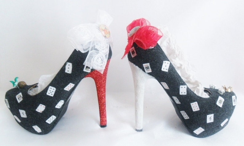 May include: A pair of black high-heeled shoes with white playing card designs. One shoe has a red glitter heel and a white bow, while the other has a white heel and a red bow.
