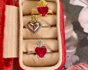 May include: Three silver and gold rings with red heart-shaped stones and ornate details are displayed in a red velvet ring box.