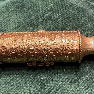 May include: A gold-colored metal container with a floral design and a hinged lid. The container has a small loop on one end, suggesting it may be a charm or pendant.