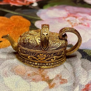 Puede incluir: Una tetera pequeña de color dorado con intrincados diseños florales. La tetera tiene un asa y un pico, y está decorada con un patrón floral.