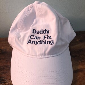 May include: White baseball cap with the text "Daddy Can Fix Anything" embroidered in blue.