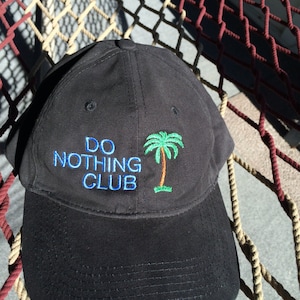 May include: A black baseball cap with a palm tree and the text "DO NOTHING CLUB" embroidered in blue and white.