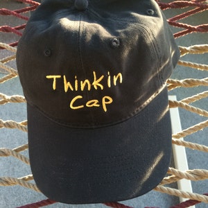 May include: A black baseball cap with a yellow embroidered design that says "Thinkin Cap".