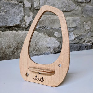 May include: A light-coloured wooden phone stand with an oval-shaped cutout and a small shelf. The word "stood" is etched on the front. Two small holes are drilled on the sides. The stand is on a white surface.